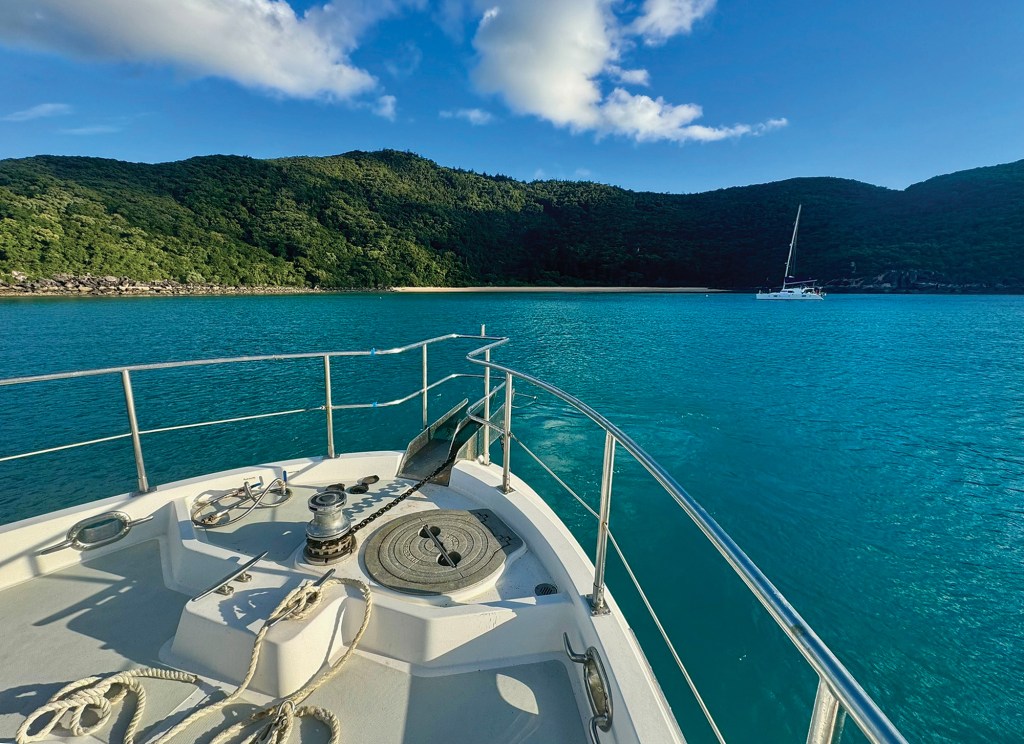 A view looking over the bow of a yacht heading through vibrant blue waters toward a lush, forested island coastline under a sunny sky with fluffy white clouds.