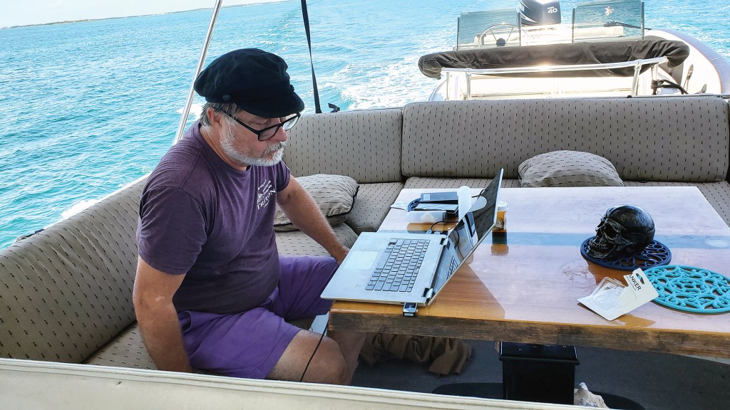 A man wearing a captain's cap working on a laptop at a table on the aft deck of a moving yacht.