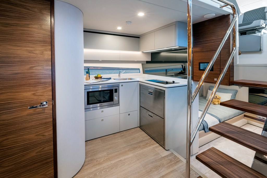 A modern lower-deck kitchen (galley) and companionway area inside a luxury yacht, showcasing sleek cabinetry and warm wood accents.

