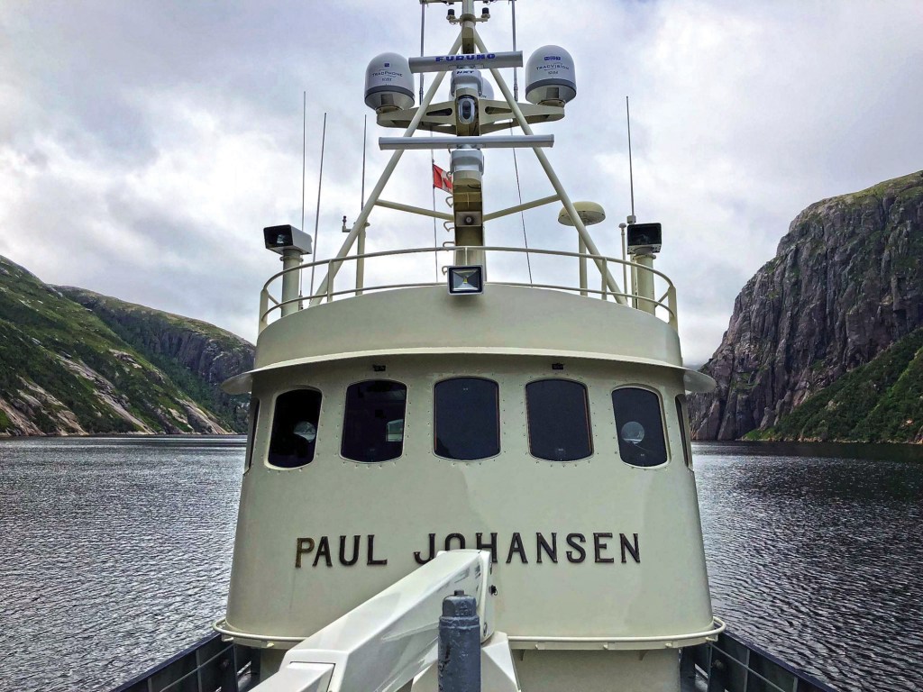 A close-up, front-facing shot looking directly at the cream-colored pilothouse of the trawler Paul Johansen as it navigates through a narrow fjord. The boat’s name, "PAUL JOHANSEN," is prominently displayed in dark metal lettering on the forward superstructure below a row of small, rounded windows. Above the bridge, a white metal mast is equipped with Furuno and KVH marine electronics, radar domes, and satellite antennas. Steep, rocky green cliffs rise up sharply on both sides of the calm water under a heavy, overcast sky.