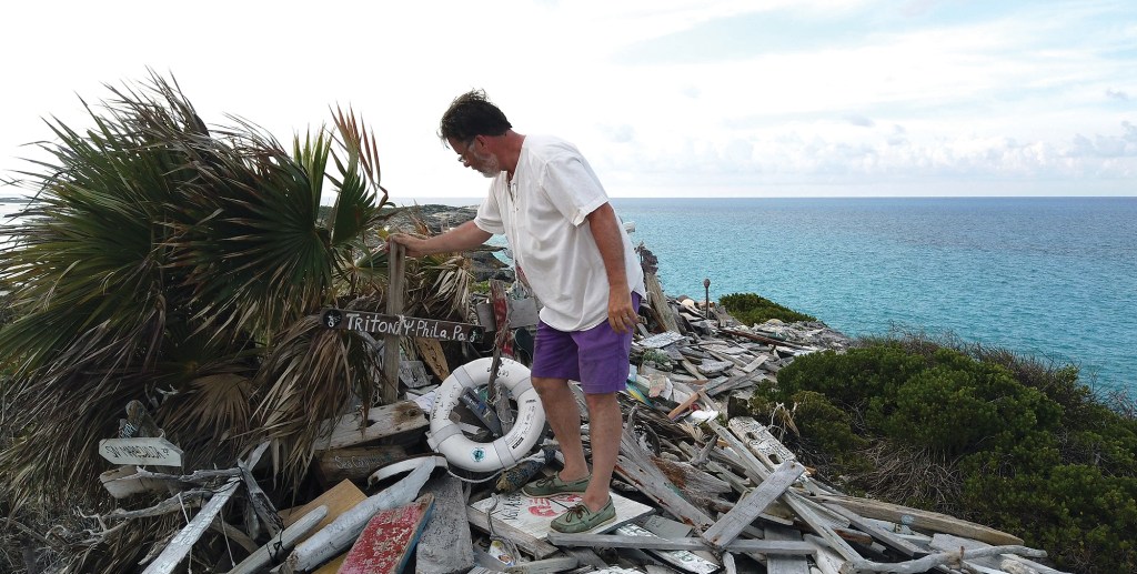 A man placing a wooden boat sign on a hilltop memorial pile made of drift wood and boat memorabilia overlooking the ocean.
