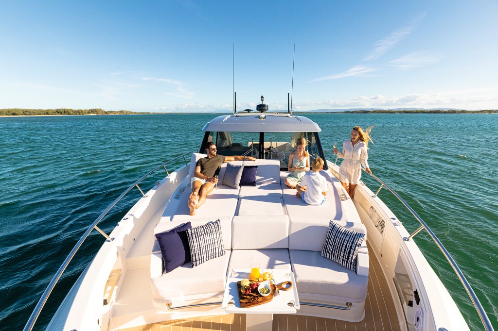 A bright, elevated bow view looking back toward the hardtop cabin of a luxury motor yacht, where a family is relaxing on the foredeck sunbed on a sunny day.
