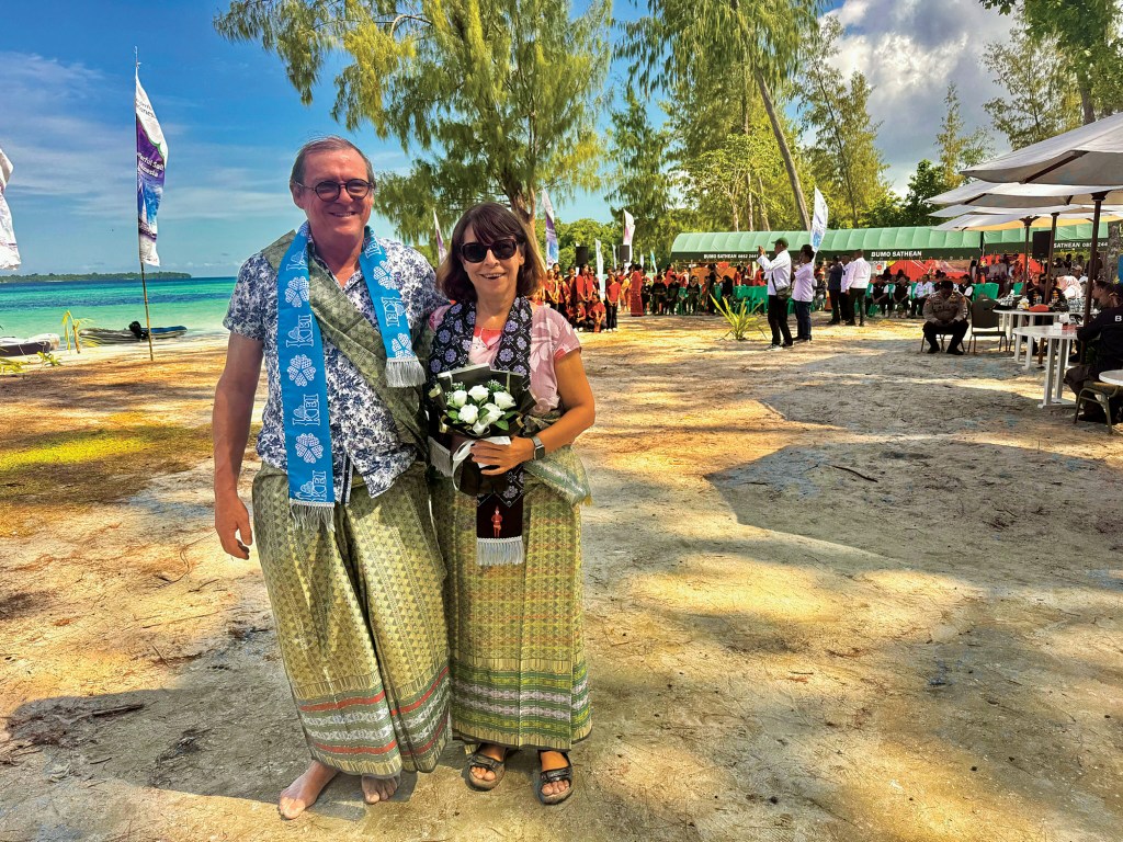 A man and woman wearing traditional patterned sarongs, ceremonial sashes, and sunglasses stand together on a sandy beach during a outdoor event by the ocean.