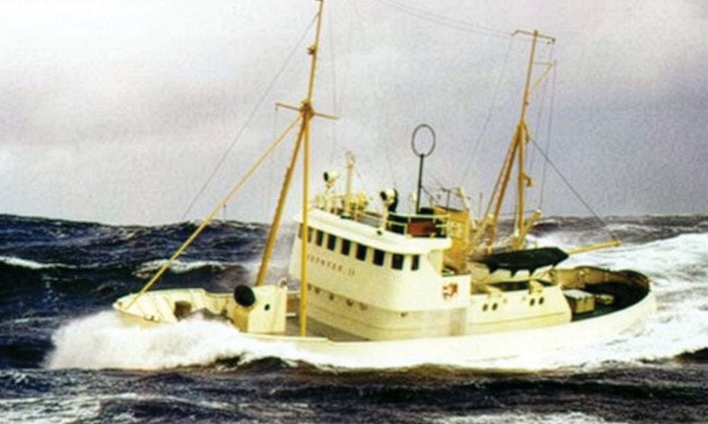 A vintage, grainy three-quarter view photo of the white rescue vessel Paul Johansen navigating rough, storm-tossed seas during its active commercial career in Norway. The boat is pitching forward, throwing up a heavy white spray of crashing foam at its bow while surrounded by deep, dark rolling waves. It features two tall yellow masts with support cables, an old-fashioned circular radio loop antenna on the upper deck, and a high-profile white pilothouse under a dramatic, overcast storm sky.