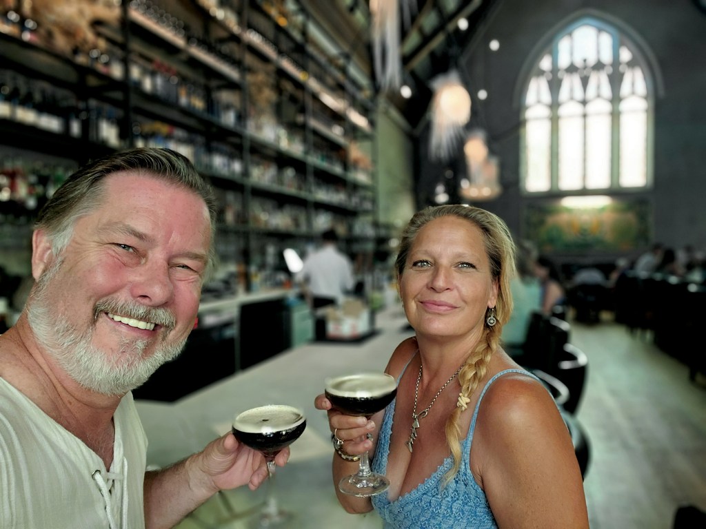 A man and a woman smiling while holding cocktail glasses inside a stylish bar with high ceilings and a large arched window.