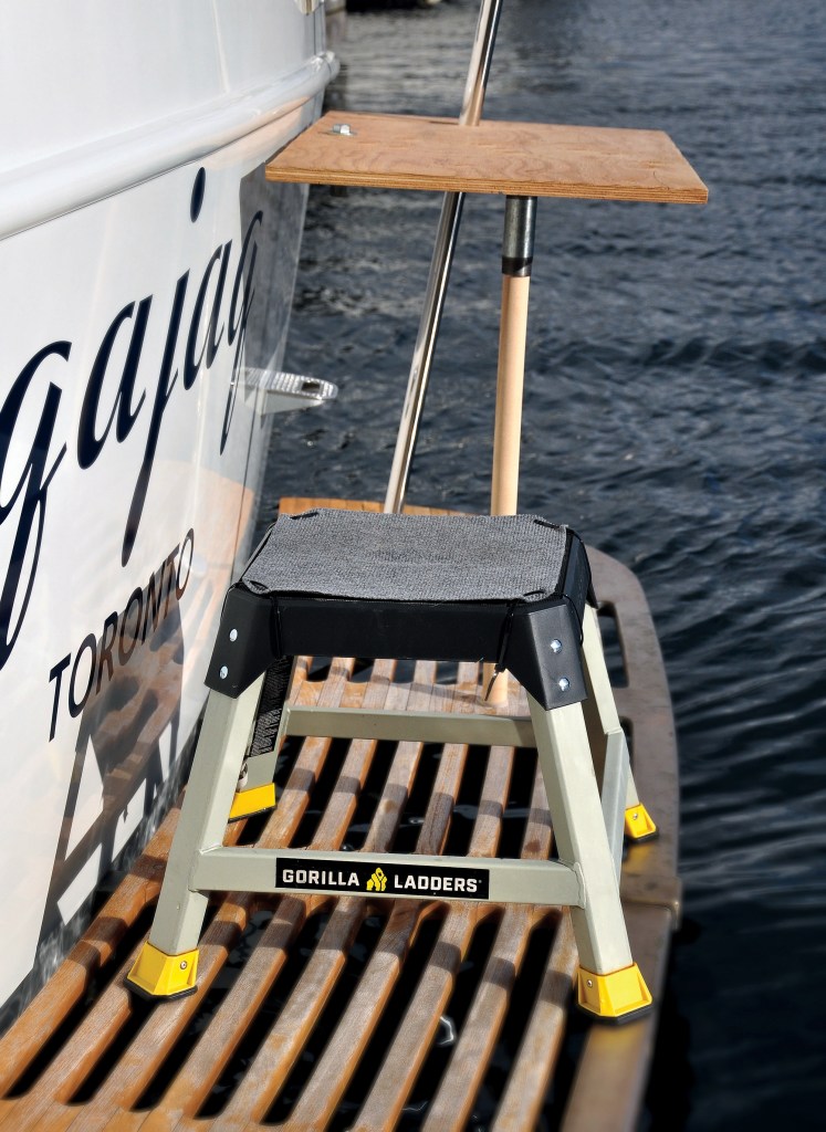 A Gorilla Ladders step stool with yellow feet sits on a teak-slatted swim platform next to a custom-built wooden boarding step mounted to the transom of a yacht.