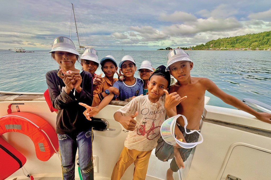 A group of smiling local children wearing silver sun visors posing together on the deck of a boat anchored near a tropical island shore.