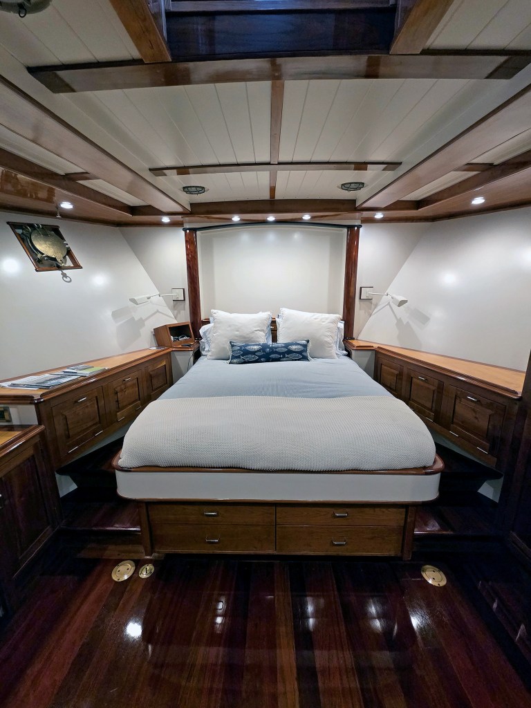 An interior view looking straight ahead at the master stateroom cabin on the trawler Paul Johansen. A large island queen bed sits at the center, neatly made with a light blue comforter, a white knit throw blanket, and a blue-and-white fish pattern lumbar pillow. Built-in, varnished wooden dressers and storage cabinets with drawers line both angled sides of the cabin wall. The room features dark polished wood flooring, a white slatted ceiling with exposed wooden beams, and soft LED accent lighting embedded above the headboard.