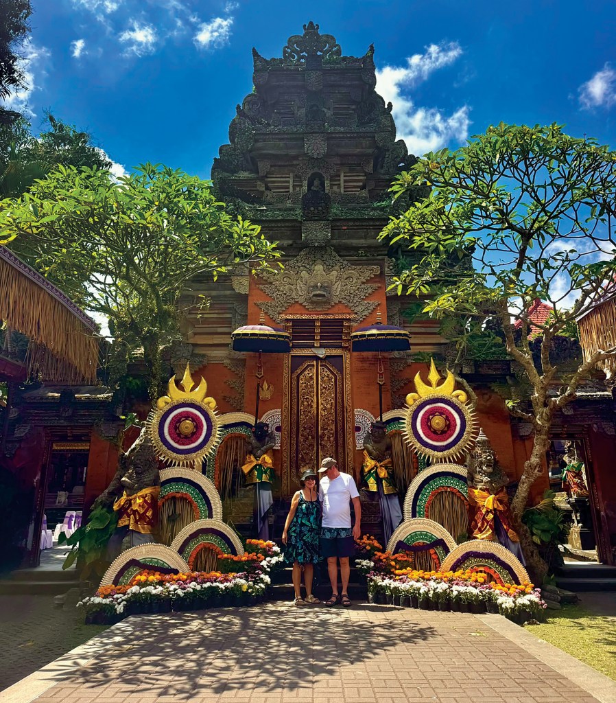 A couple stands together in front of an intricately carved Balinese temple structure, decorated with colorful floral arrangements and ceremonial umbrellas at Ubud Palace under a bright blue sky.