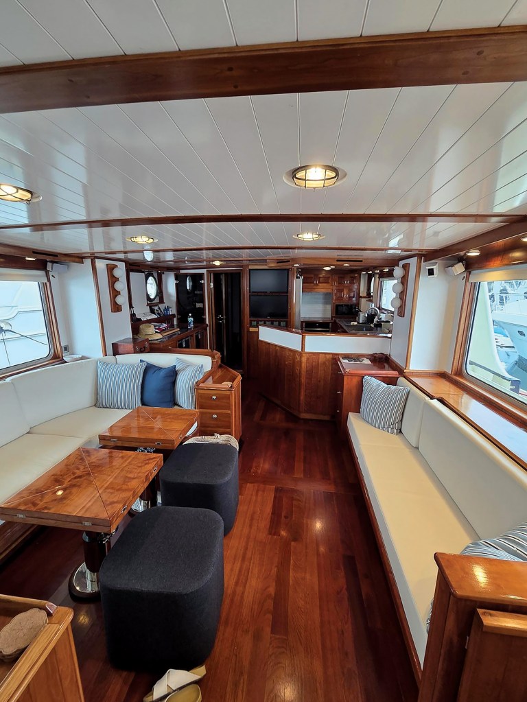 An interior view looking aft down the long, open-plan salon and galley of the trawler Paul Johansen. The living area features white built-in sofas with light cream cushions and striped accent pillows lining both sides of the cabin. On the left, two high-gloss burl wood coffee tables are paired with two dark grey fabric storage ottomans over a polished dark wood floor. In the background, a varnished wood-paneled breakfast bar separates the salon from the galley kitchen. The room is brightly lit by large side windows and recessed ceiling lights set into a white slatted overhead with exposed wood beams.