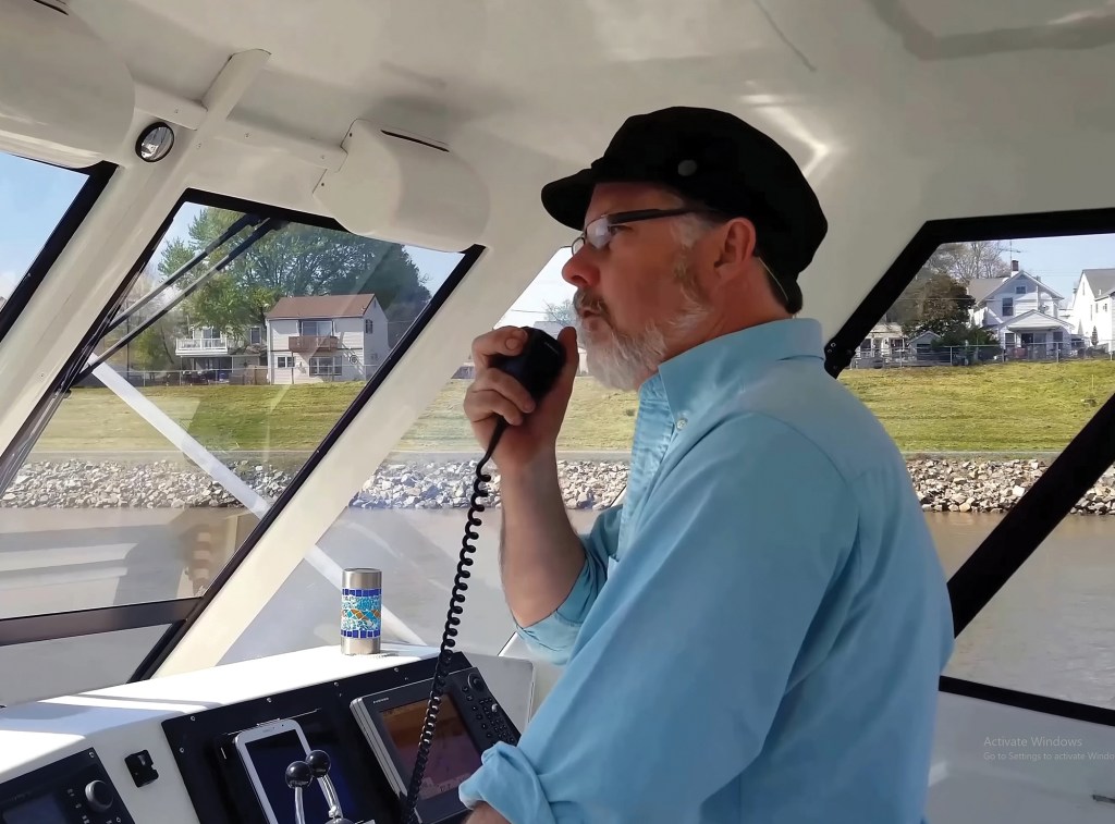 A boat captain wearing glasses and a cap speaking into a VHF marine radio microphone at the helm.