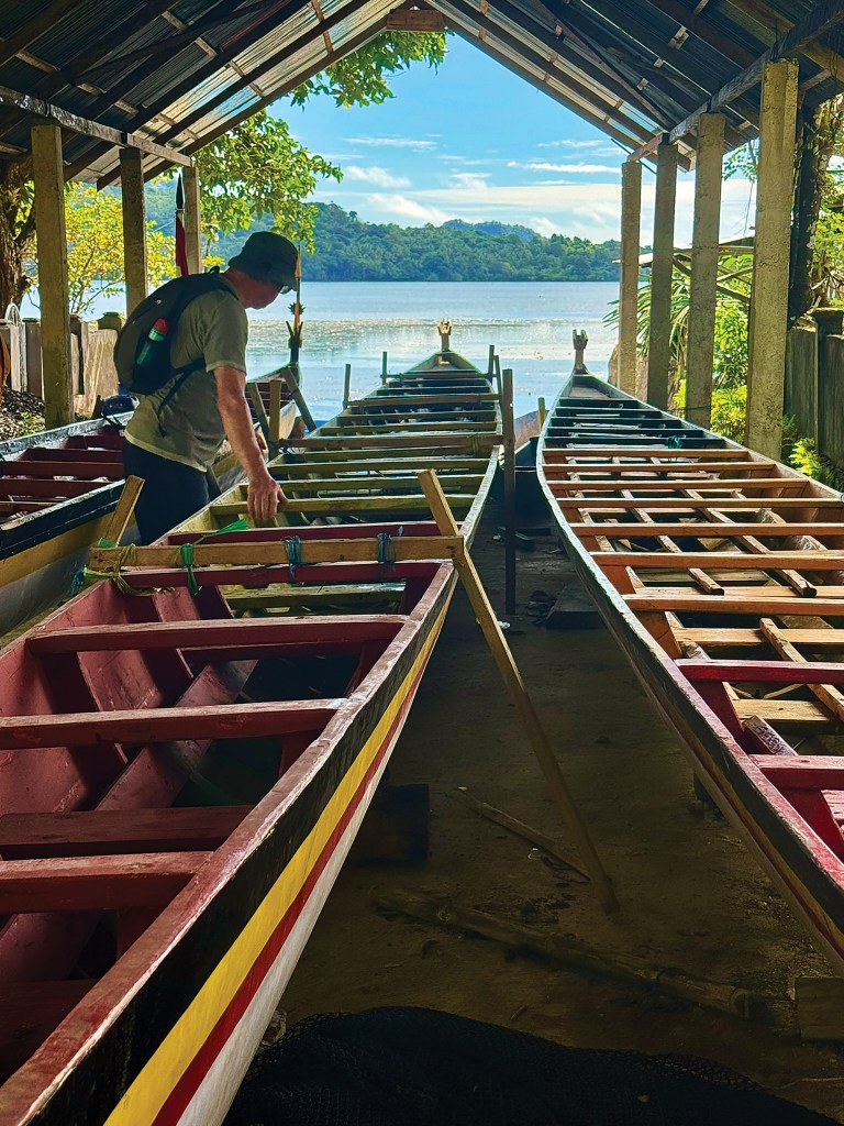 A man inspects traditional long wooden war canoes stored under a covered open-air boat shed overlooking a tropical bay.