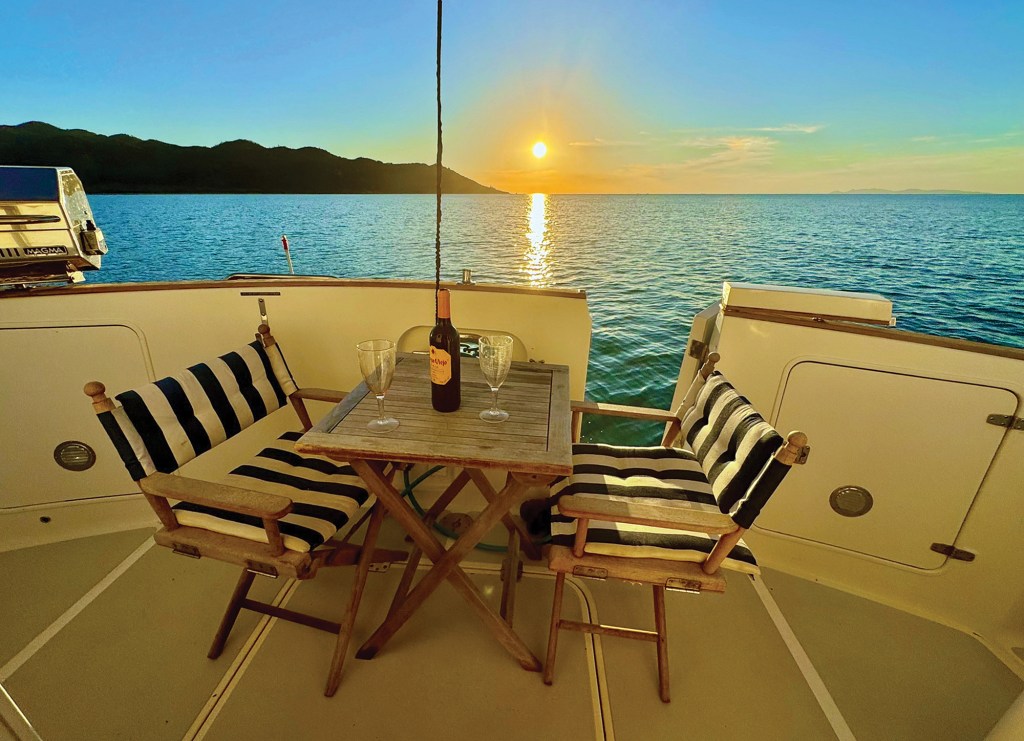 A wooden table set with two wine glasses and a bottle of wine between two striped folding chairs on a boat deck, overlooking a peaceful sunset over the ocean in Florence Bay, Australia.