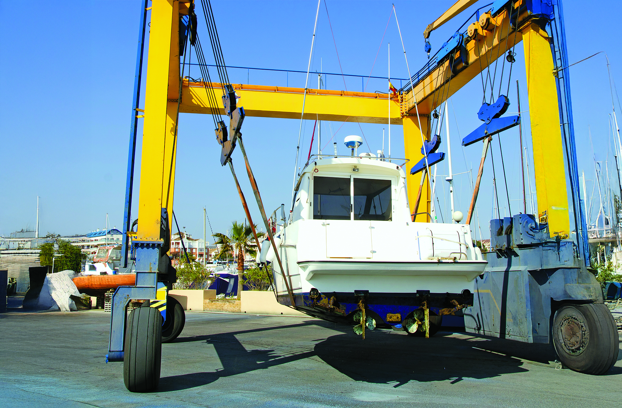 A white motorboat suspended in lifting straps on a large yellow gantry travel lift at a boatyard or marina on a clear day.