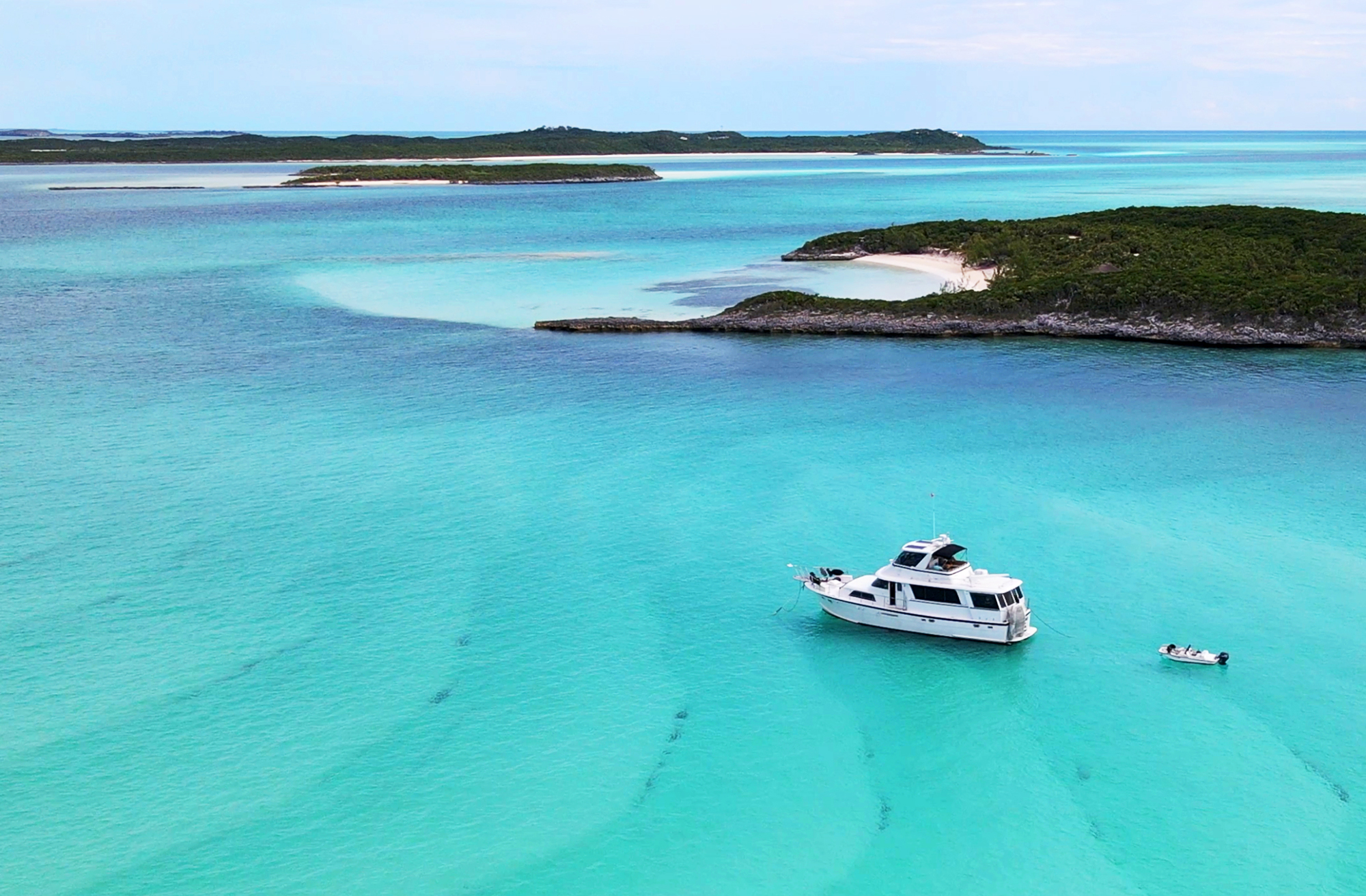 An aerial view of a white motor yacht anchored in clear turquoise tropical water near lush green islands with sandy beaches.