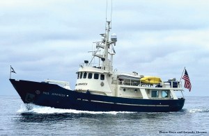 A side three-quarter view of a large, rugged expedition trawler named "PAUL JOHANSEN" cruising through calm open water. The vessel features a dark blue hull creating a white wake at the bow, paired with a cream-colored superstructure and a high pilothouse. A tall central mast holds multiple radar domes and communication antennas, while the upper deck carries a white tender and a bright yellow kayak. An American flag flies from the stern under a grey, overcast sky.