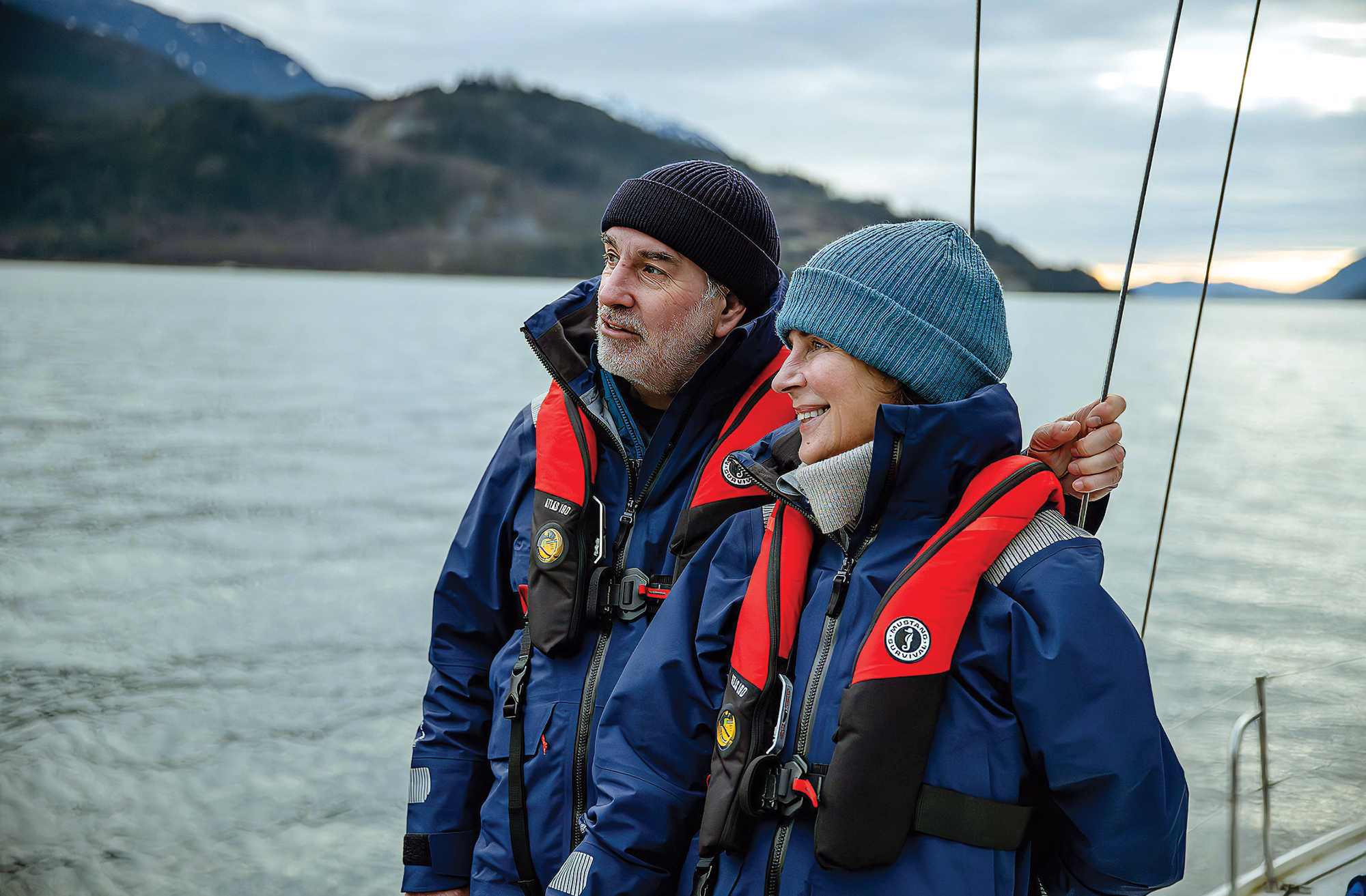 A man and woman standing together aboard a boat on a cold, overcast day, wearing matching blue weather coats, beanies, and red-and-black Mustang Survival inflatable PFDs.