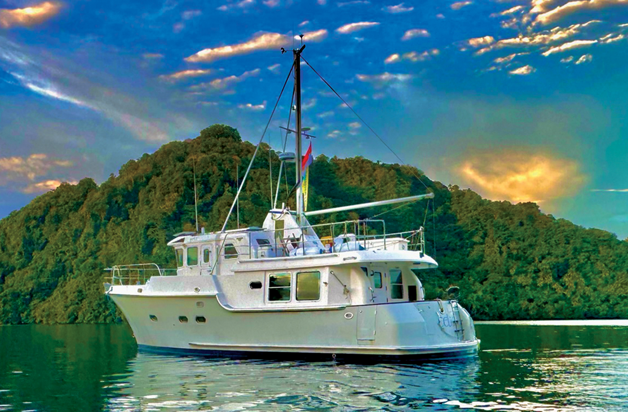 A white Nordhavn 43 expedition yacht anchored in calm water near a lush green island with a dramatic, vibrant sky filled with glowing clouds overhead.