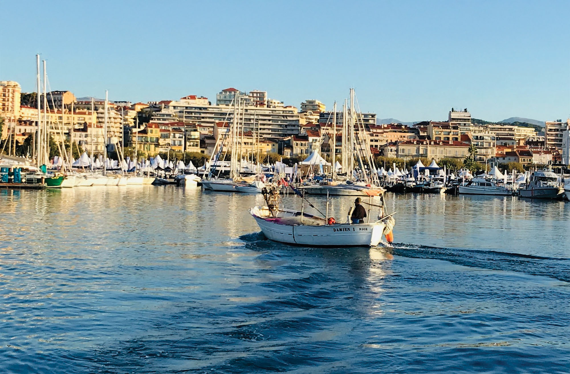 A wide, eye-level shot of a small, white wooden fishing boat named "DAMIEN I NICE" cruising through a calm harbor, leaving a gentle wake in the foreground. A single fisherman sits at the stern near the outboard tiller. In the background, a bustling marina is packed with dozens of docked sailboats and white vendor tents along the waterfront, bordered by a dense hillside of European-style apartment buildings under a clear blue sky.