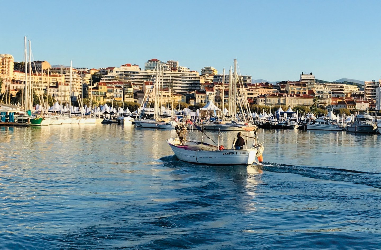A wide, eye-level shot of a small, white wooden fishing boat named "DAMIEN I NICE" cruising through a calm harbor, leaving a gentle wake in the foreground. A single fisherman sits at the stern near the outboard tiller. In the background, a bustling marina is packed with dozens of docked sailboats and white vendor tents along the waterfront, bordered by a dense hillside of European-style apartment buildings under a clear blue sky.