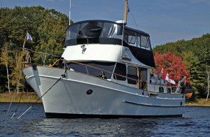 feat_dog-on-board A classic white flybridge trawler yacht anchored in a calm body of water with a dark-furred dog peeking over the bow railing, set against a background of colorful autumn trees.
