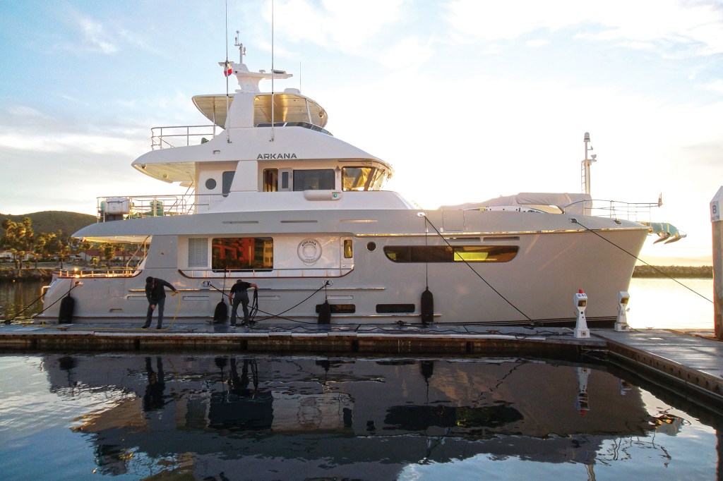 The Nordhavn 71 expedition yacht Arkana docked alongside a marina pier, illuminated by the warm light of sunrise as crew members handle dock lines.