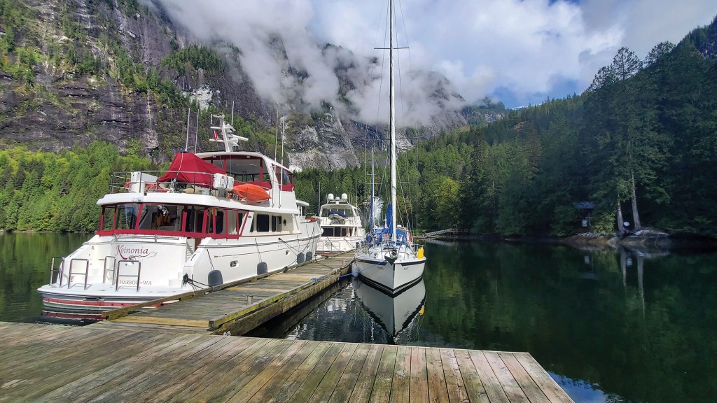 A motor yacht named "Koinonia" and a sailboat moored alongside a wooden dock in calm waters, set against a backdrop of steep, cloud-capped granite cliffs and dense pine forest in Princess Louisa Inlet.