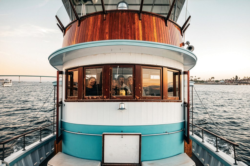 Close-up front view of the wheelhouse on the classic wooden yacht Dickie Walker, showing passengers smiling through the wood-framed windows, rich varnish detailing, and blue-and-white painted paneling while cruising on open water.