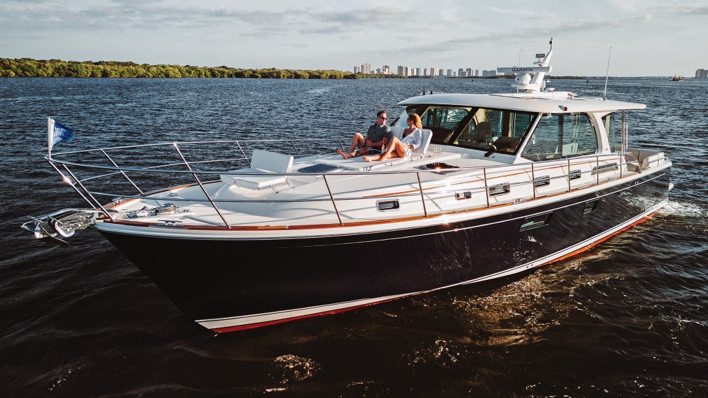 A dark-hulled Sabre 51 Salon Express cruising across open water in late-afternoon sun, with a man and woman relaxing on the bow sunpad facing the shore skyline.