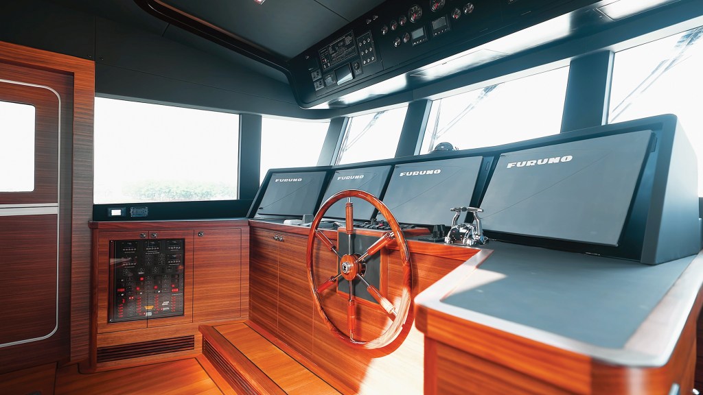 The pilothouse helm station of a Selene 64 Ocean Explorer yacht, featuring a classic wooden Destroyer wheel, four large Furuno displays, dark American walnut wood paneling, and an overhead console.