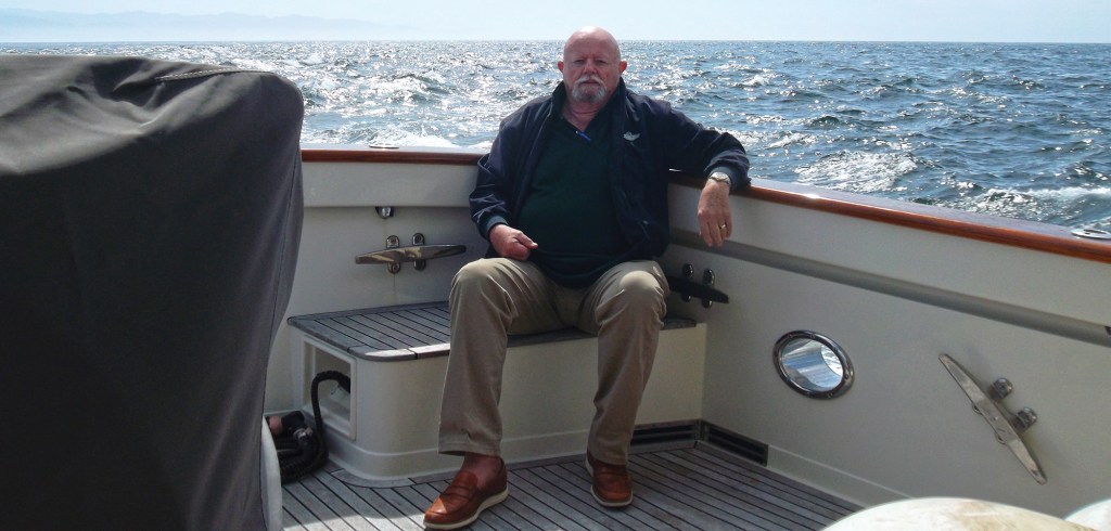 A man sitting on the aft deck bench of a Fleming 65 motor yacht while underway on open, sparkling waters.