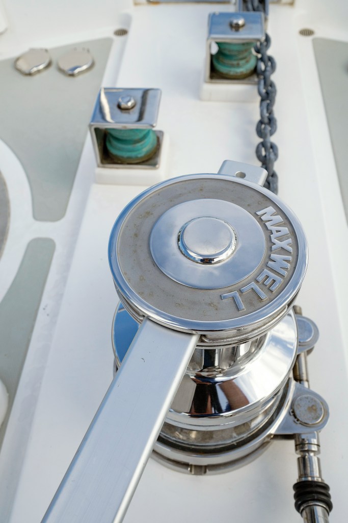Top-down view of a stainless-steel Maxwell windlass with a manual clutch handle attached and an anchor chain extending along the foredeck.