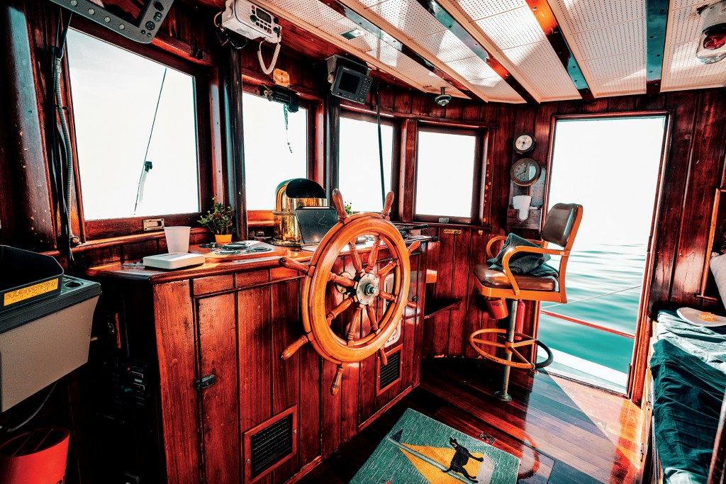 Interior view of the restored wooden wheelhouse on the Dickie Walker, featuring a classic spoked wooden ship's wheel, rich polished timber panelling, brass instruments, helm chair, and large windows looking out over calm water.