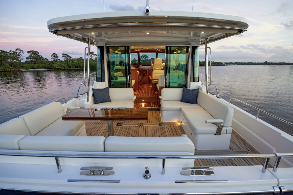 A view looking forward from the aft deck into the open-air cockpit and main salon of a Sabre 51 Salon Express, showcasing teak decking, plush white L-shaped seating, a polished wooden table, and glass sliding doors leading into the illuminated interior.