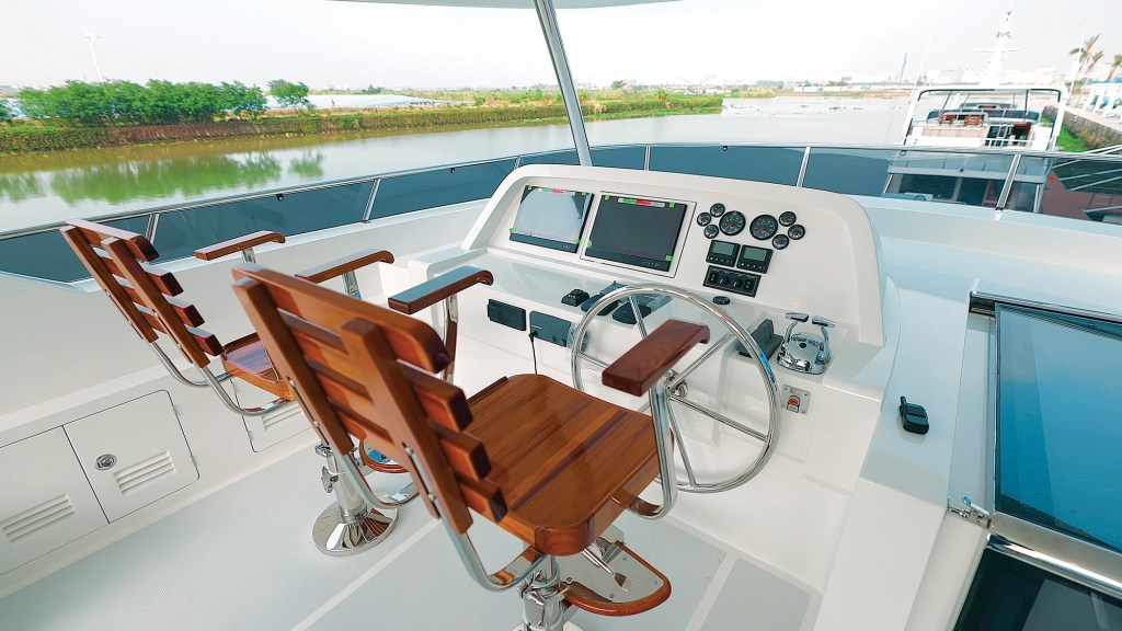 The flybridge helm station of a Selene 64 Ocean Explorer yacht, featuring two polished wooden helm chairs, a stainless steel steering wheel, dual navigation screens, and engine gauges.