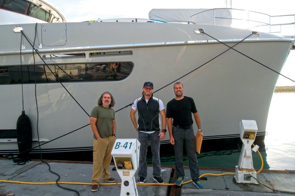 Three crew members pose on a concrete dock beside slip B-41 in front of the hull of a grey Nordhavn 71 yacht.