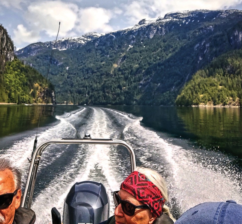 A view looking sternward from a small powerboat leaving a foamy wake across calm water, with two passengers in sunglasses in the foreground and a backdrop of steep, pine-covered mountains under a partly cloudy sky.