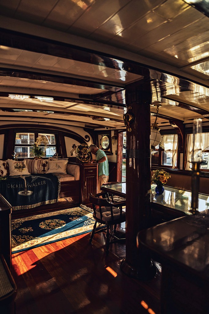 Warmly lit luxury salon inside the classic wooden yacht Dickie Walker, featuring Brazilian cherry wood floors, polished brass accents, built-in daybed seating with custom embroidered throw pillows, an ornate dining area, and an antique bronze dive helmet.