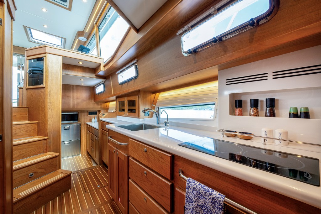 The galley-down area of a Sabre 51 Salon Express featuring rich cherrywood cabinetry, solid-surface countertops with a electric cooktop and sink, teak-and-holly flooring, and stairs leading up to the main salon.
