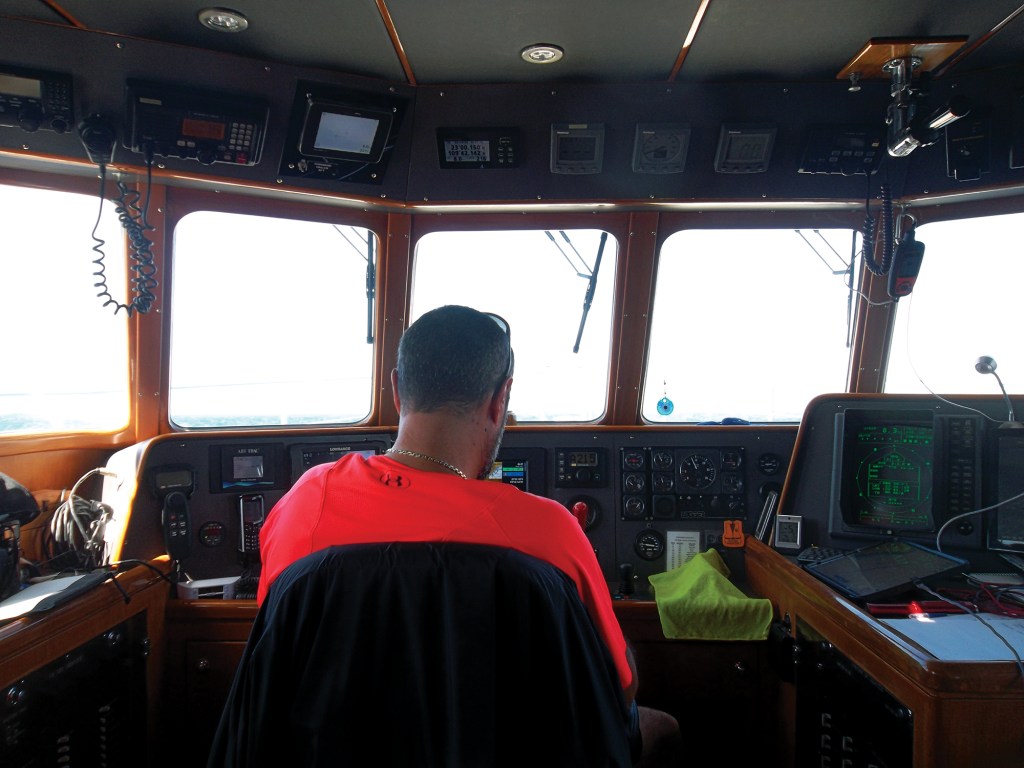 A captain in a bright red shirt sitting at the helm station inside the pilothouse of a Nordhavn 50 yacht, surrounded by navigation displays, instruments, and large forward-facing windows looking out to open sea.