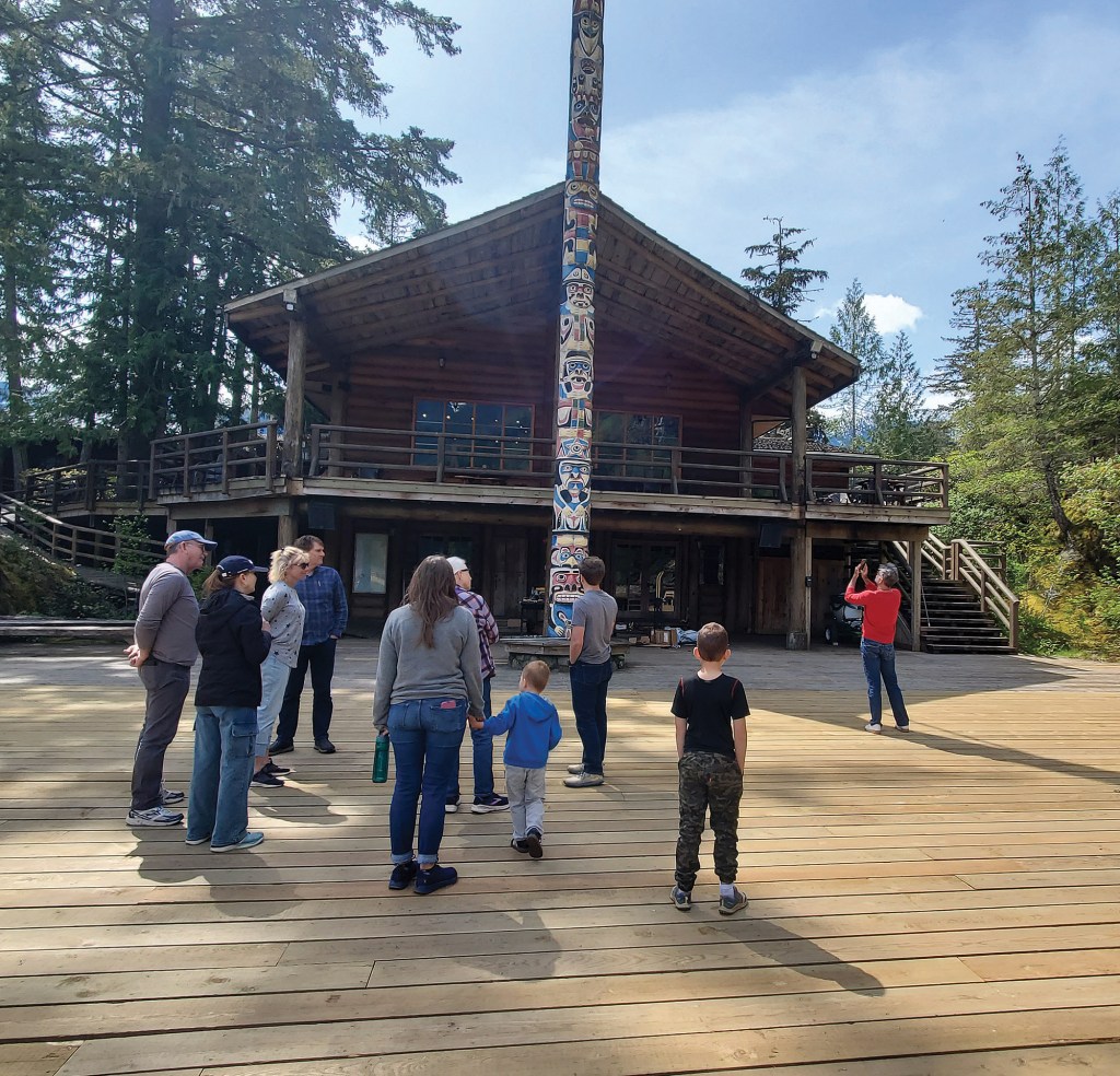 A group of visitors gathered on a spacious wooden deck outside a two-story heavy-timber lodge, looking up at a tall, intricately carved Indigenous totem pole extending past the building's roofline.