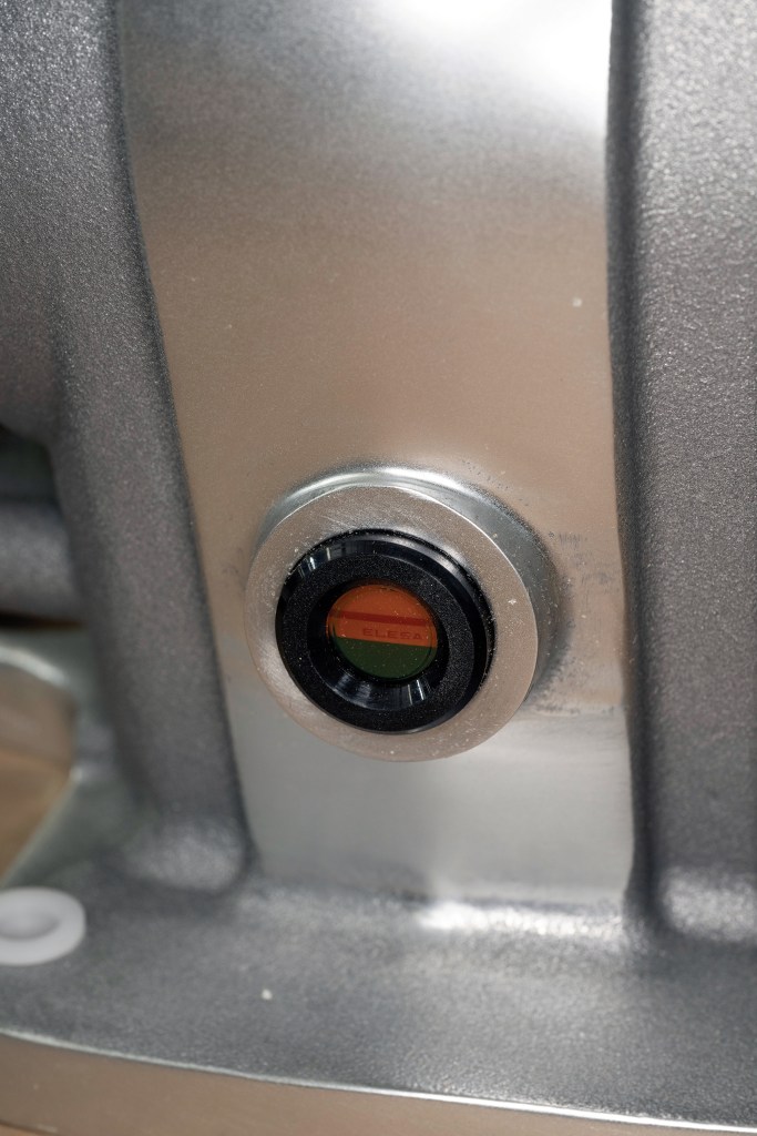 Extreme close-up of an oil level sight glass with red and green indicator lines on a metallic windlass gearbox housing.