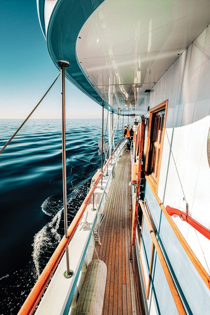 Looking down the side deck of the classic wooden yacht Dickie Walker while underway, showing teak decking, varnished handrails, signature red-painted fire axe mounted on white paneling, and calm blue ocean water.