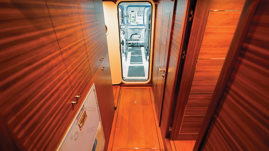 A walnut-paneled hallway inside a Selene 64 Ocean Explorer yacht, looking through an open watertight door directly into the brightly lit engine room.