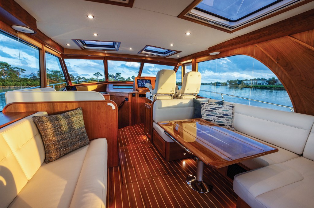 The main salon of a Sabre 51 Salon Express filled with warm golden sunlight, showcasing cream leather seating, a high-gloss dining table, two helm chairs, and large surrounding windows with panoramic views.