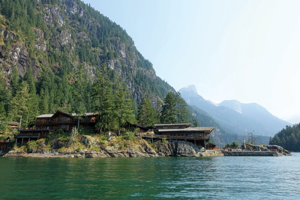 A wide shot taken from the water showing a rustic, heavy-timber lodge resort complex perched along the rocky, tree-lined shore of a fjord, with steep, evergreen-covered mountains sloping upward in the background under a bright sky.