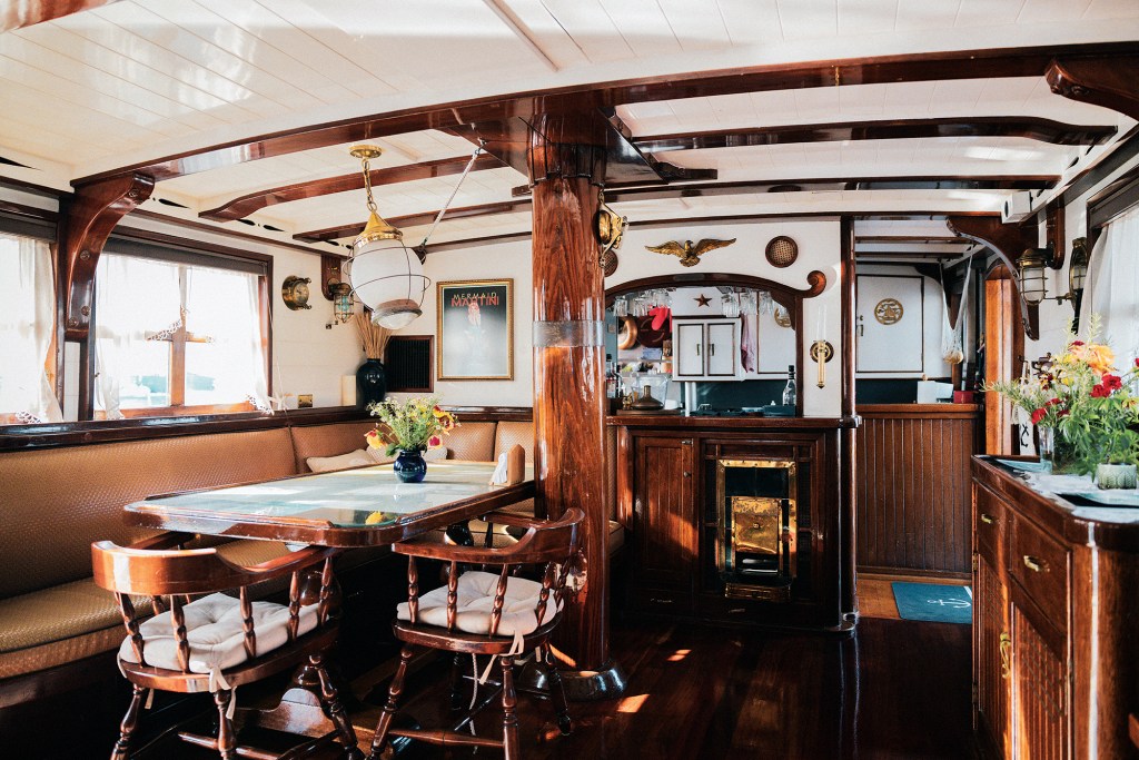 Interior view of the main dining area and galley inside the classic wooden yacht Dickie Walker, featuring a wooden table with banquette seating, a polished wooden support pillar, overhead maritime lighting, framed nautical art, and rich cherry wood cabinetry.