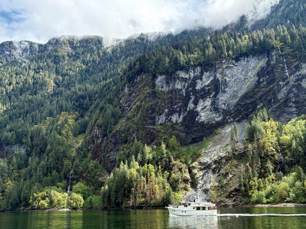 A white motor yacht cruising along dark, calm fjord waters, dwarfed by towering granite cliffs and dense, evergreen-forested mountains with low-hanging clouds misting over the peaks in Princess Louisa Inlet.