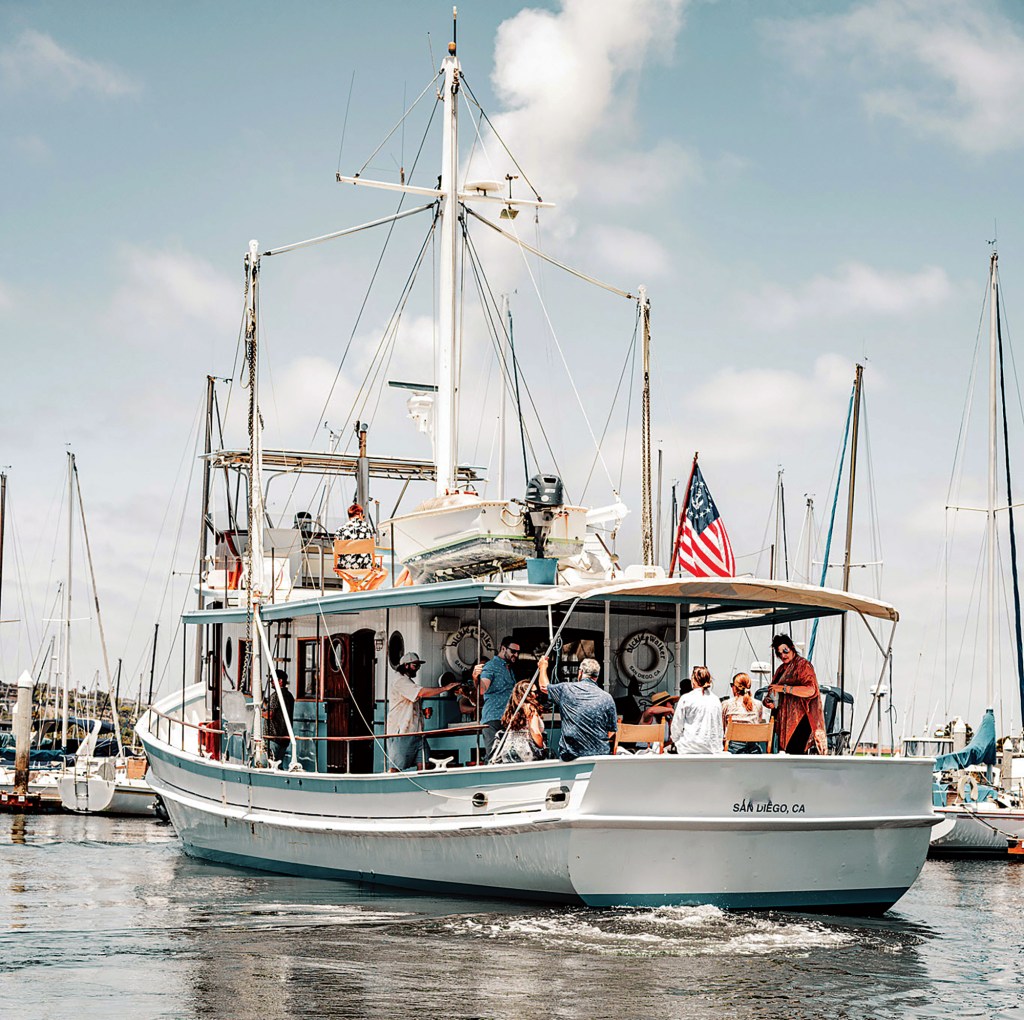 A stern view of the classic wooden yacht Dickie Walker docked in a harbor, showing guests gathered on the aft deck beneath a sunshade, an American flag flying, a tender mounted on the upper deck, and "San Diego, CA" painted across the transom.