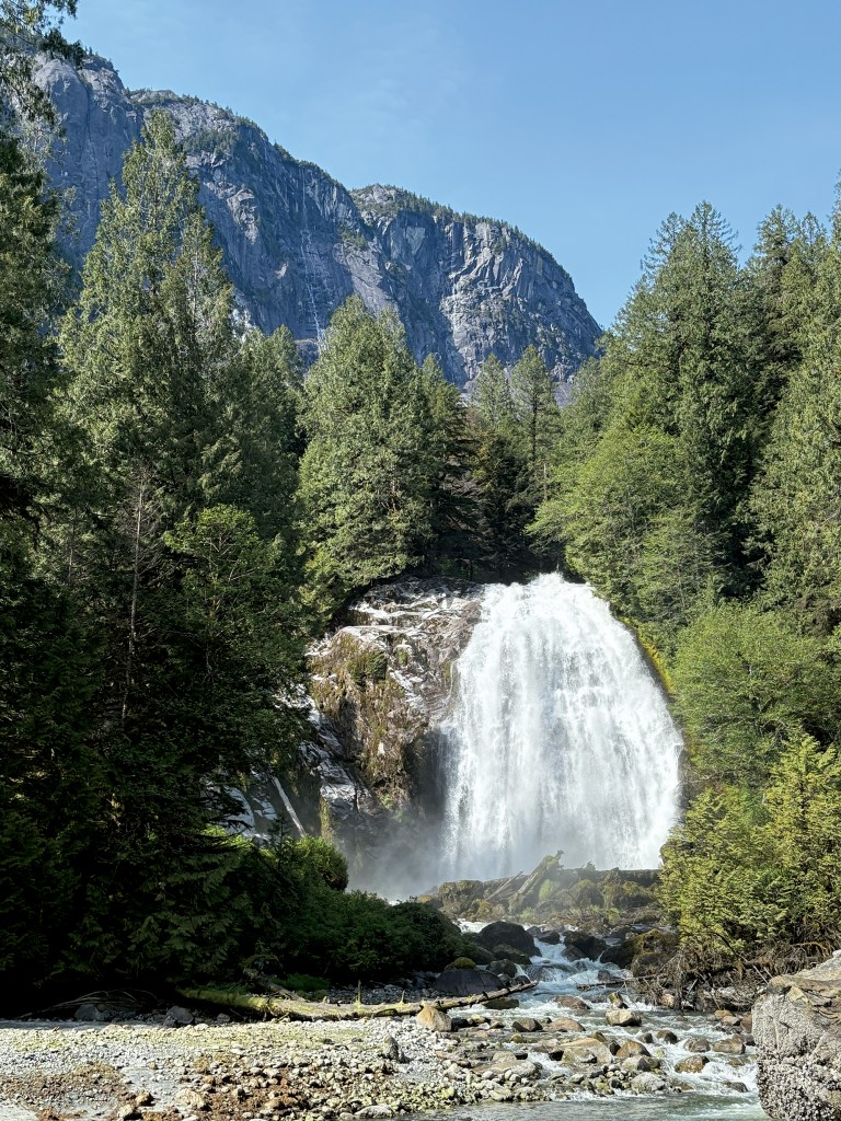 A dramatic vertical view of Chatterbox Falls cascading powerfully over rocky rock faces into a rocky riverbed, framed by dense green evergreen trees with sheer, towering granite peaks rising under a clear blue sky in the background.
